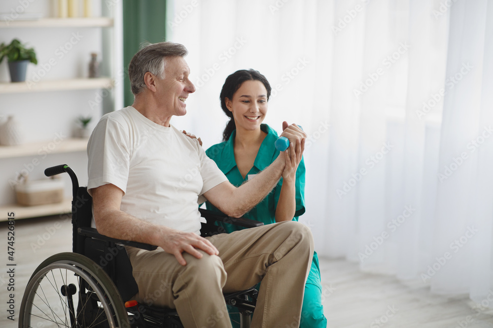 Young female physiotherapist helping senior man in wheelchair to work out with dumbbells, to recover from injury Young female physiotherapist helping senior man in wheelchair to work out with dumbbells, to recover from injury