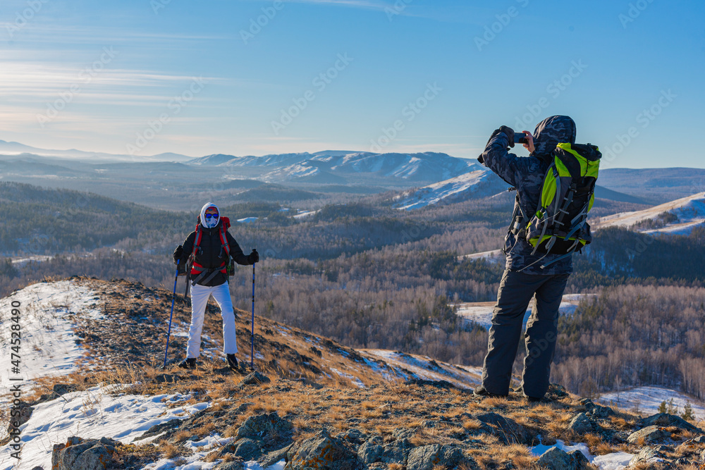 Middle-aged man takes a photo of his wife with a smartphone while hiking in the winter alpine mountains at sunrise.