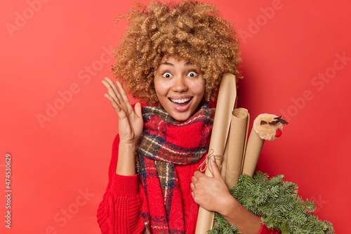 Optimistic woman with curly hair raises palm feels excited holds spruce wreath and rolled paper feels glad to celebrate Christmas holidays isolated over red background. Winter time and celebration