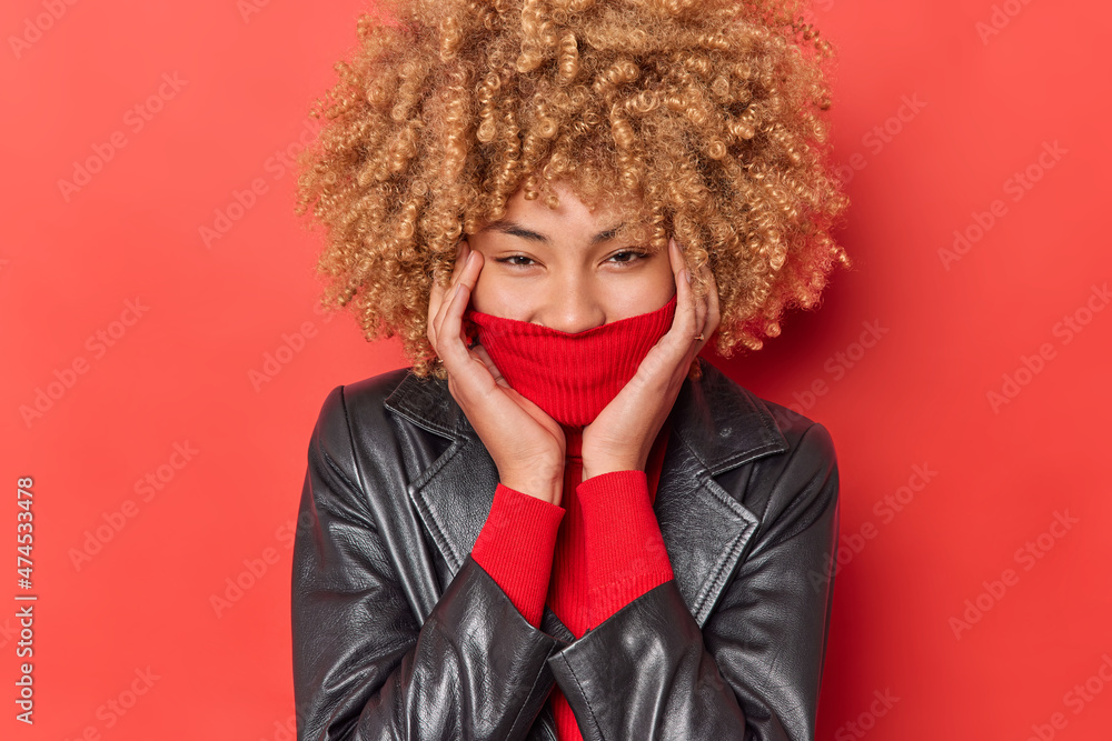 Happy positive woman hides mouth behind collar dressed in leather jacket feels carefree and joyful poses against red background has eyes full of happiness. People clothes and emotions concept