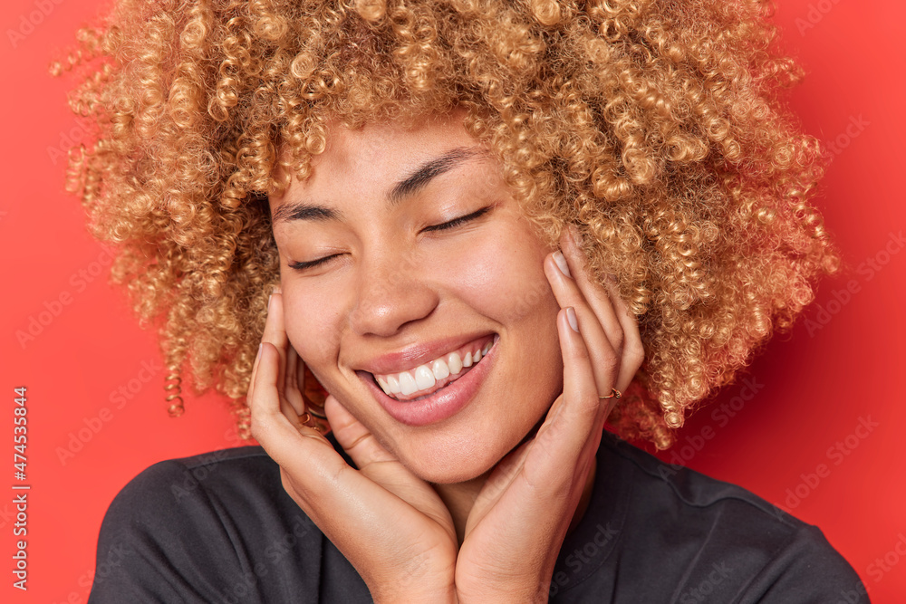 Headshot of happy woman keeps hands on cheeks closes eyes smiles toothily recalls nice memories wears casual clothes has blonde cury hair isolated over vivid red background. Positive emotions Headshot of happy woman keeps hands on cheeks closes eyes smiles toothily recalls nice memories wears casual clothes has blonde cury hair isolated over vivid red background. Positive emotions