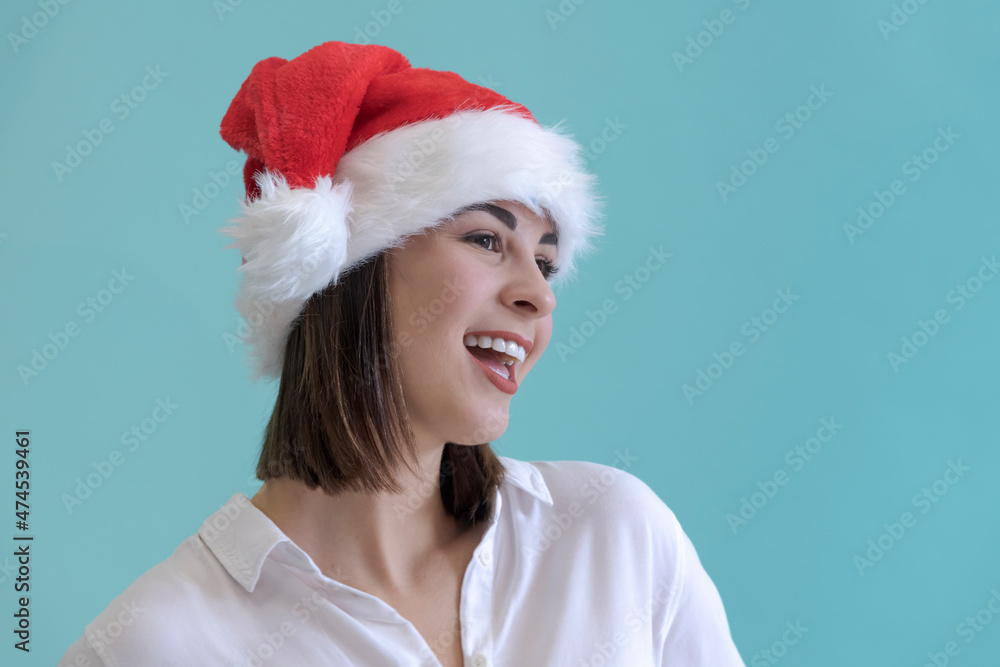 Portrait of beautiful smiling caucasian woman with brown hair wearing white shirt and red Santa Claus hat and looking away. Blue background. Christmas holidays theme. Portrait of beautiful smiling caucasian woman with brown hair wearing white shirt and red Santa Claus hat and looking away. Blue background. Christmas holidays theme.