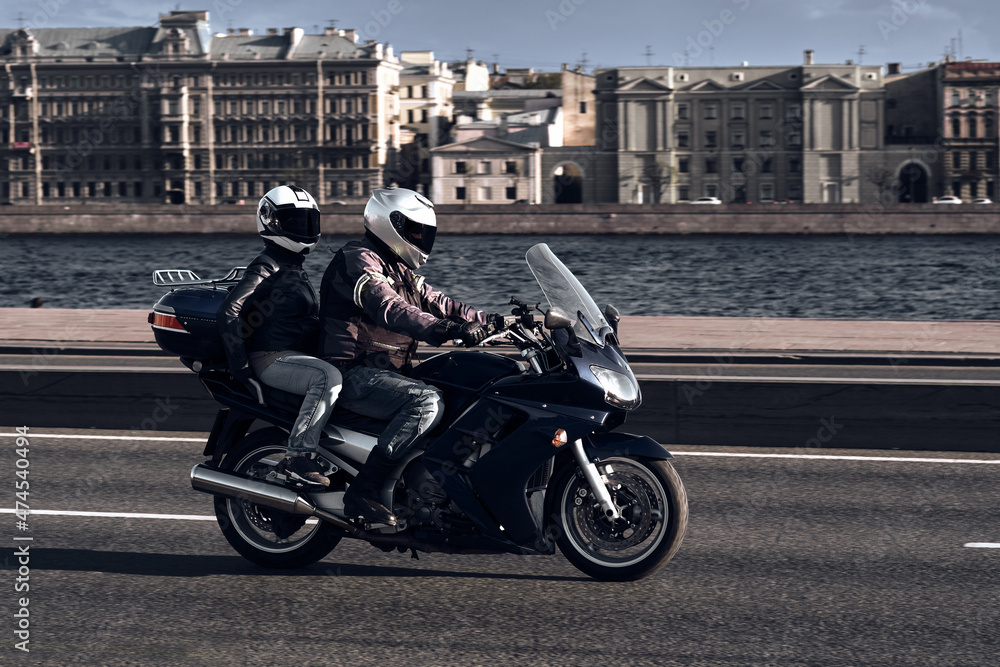 A man and a woman ride together on a sports motorcycle along the embankment. Motorcyclists drive around the city in the evening on a dark blue sports bike. A man and a woman ride together on a sports motorcycle along the embankment. Motorcyclists drive around the city in the evening on a dark blue sports bike.