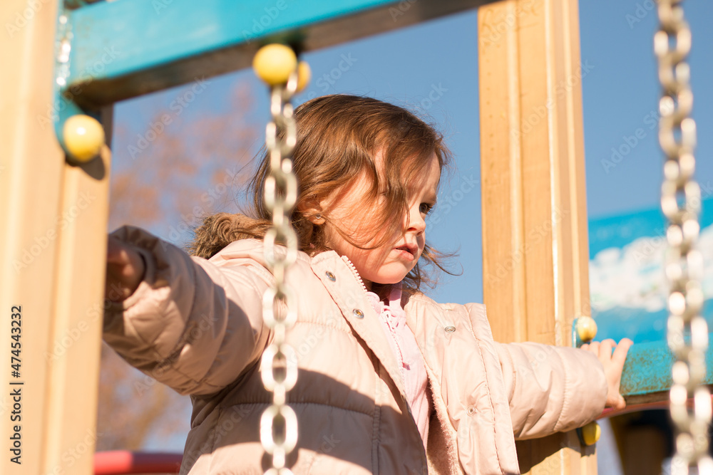 Obraz premium Small child with long red hair against the background of a blue sky in warm clothes on the playground