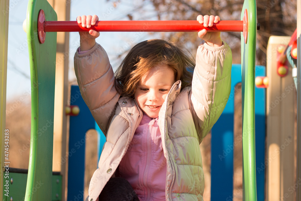 Fototapeta premium Small child with long red hair against the background of a blue sky in warm clothes on the playground