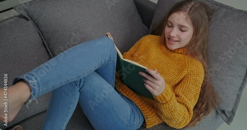 Closeup portrait cheerful student girl reading book, lying on the sofa