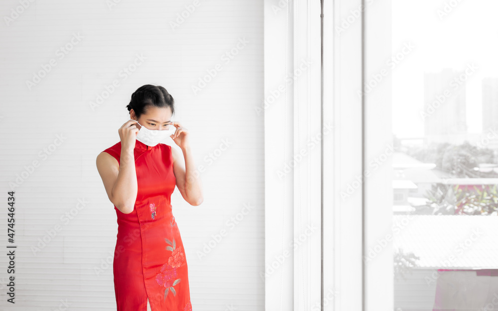 Portrait beautiful Asian woman wearing red qipao or dress, mask to protect virus, standing beside window with copy space, celebrating Chinese New Year. Religion, Healthcare and Festive concept.