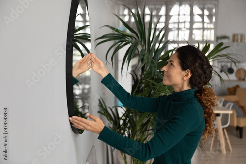 Smiling millennial Latino woman put mirror on wall decorate cozy living room move in new house or apartment. Happy young Hispanic female renovate settle in own home. Renovation, design concept. photo