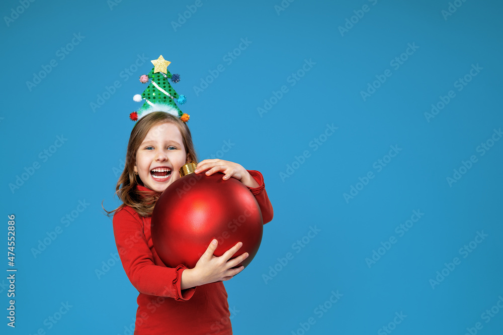 happy little girl with decorated headband holds Christmas red balloon on blue background in studio. child with Christmas tree on his head smiles happily and receives gift for Christmas. Advertising. happy little girl with decorated headband holds Christmas red balloon on blue background in studio. child with Christmas tree on his head smiles happily and receives gift for Christmas. Advertising.