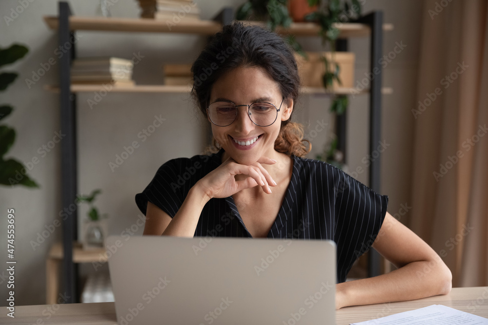Smiling young Latino woman in glasses look at laptop screen work online on  gadget at home