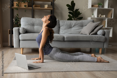 Fototapeta Sporty young Latino woman stretch on floor at home practice yoga or pilates watching video lesson on laptop