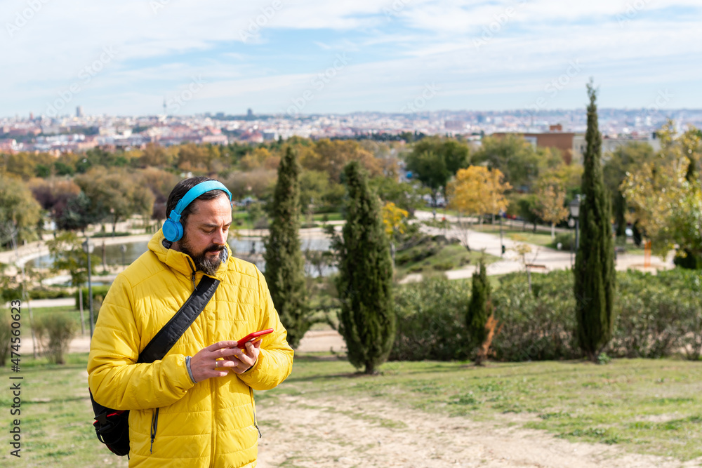 young hipster man with a beard walks through a city park in a yellow coat listening to music with wireless headphones and his smart phone