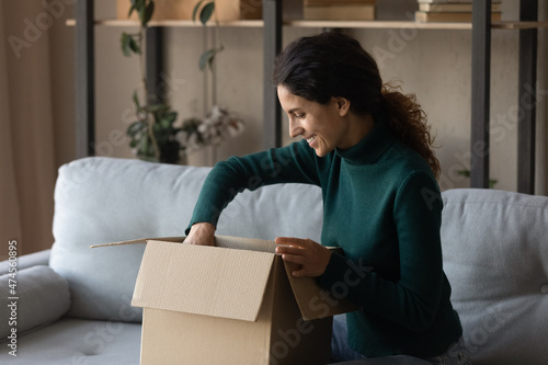 Excited millennial Hispanic female buyer or client open unbox package shopping online from home. Smiling young Latino woman unpack box with order buy on internet, satisfied with good delivery service.