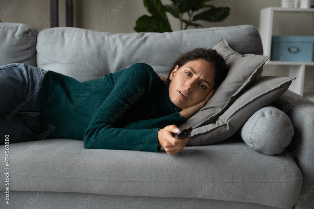 Calm young Hispanic woman lying relaxing on couch in living room watch TV switch channels with remote controller. Millennial latin female rest on sofa at home enjoy television. Entertainment concept.