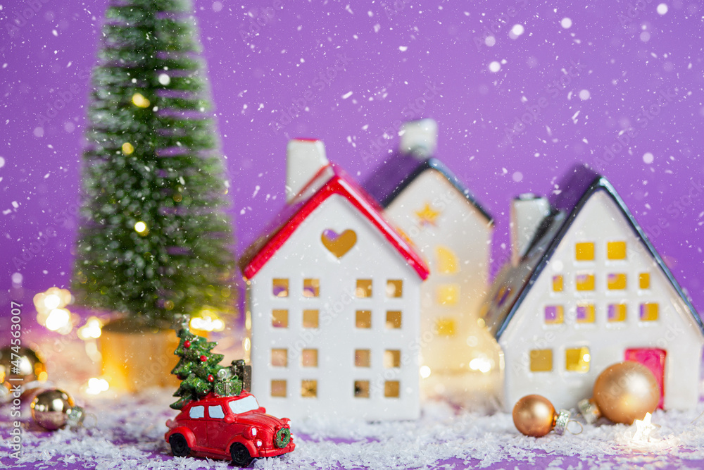 Christmas decor - red retro car on snow carries past houses with fairy lights in bokeh Christmas tree with gift boxes on roof. Toy on violet background. New Year greeting card. Cozy home