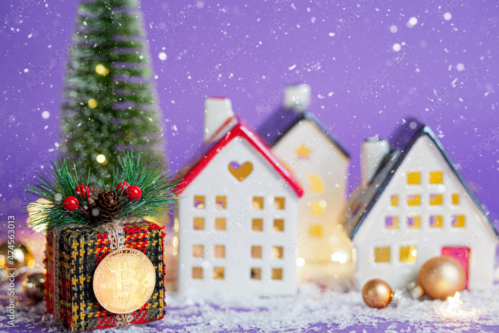 Bitcoin coin on gift box past houses with fairy lights and snow, Christmas tree. Violet background very peri. New Year greeting card. Cryptocurrency, finance, wealth and investing.