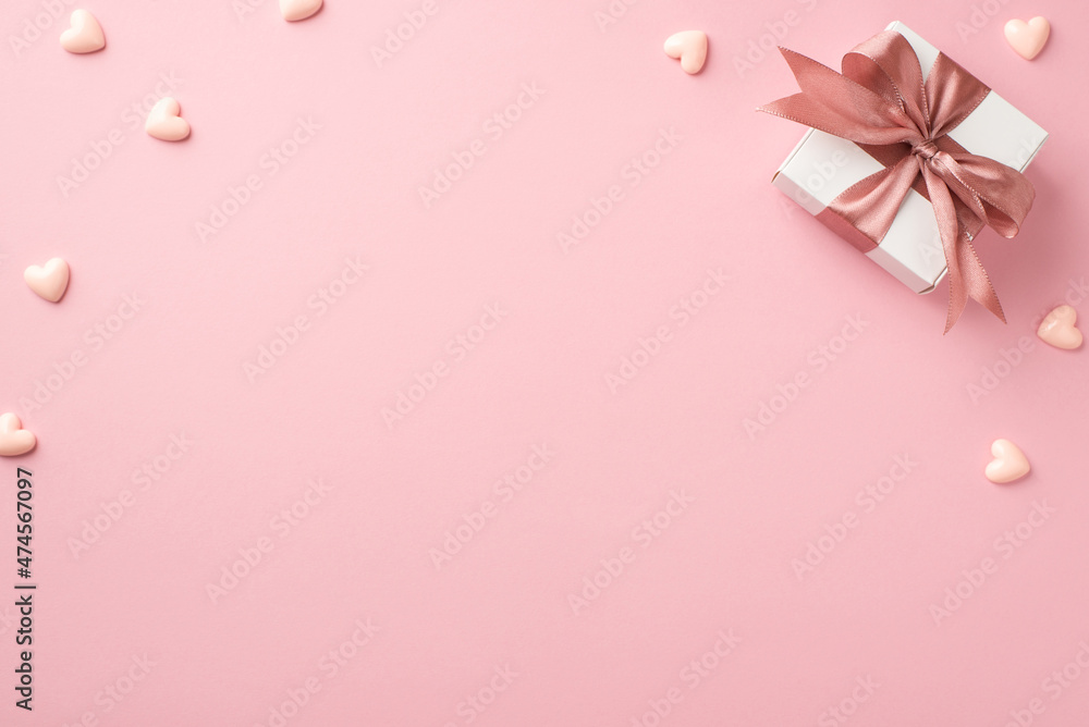 Top view photo of valentine's day decorations white giftbox with pink satin ribbon bow and small hearts on isolated pastel pink background with empty space
