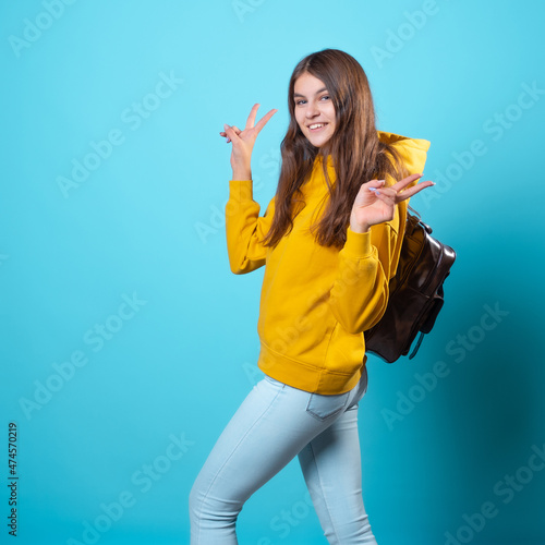 Cute young brunette in jeans and yellow hoodie. smiling student in glasses with a backpack on her shoulders holds a takeaway coffee in her hands  and shows a two thumbs up gesture