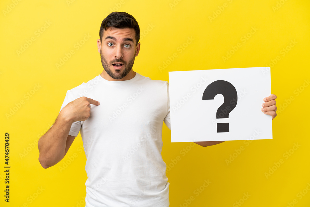 Young handsome caucasian man isolated on yellow background holding a placard with question mark symbol with surprised expression Young handsome caucasian man isolated on yellow background holding a placard with question mark symbol with surprised expression