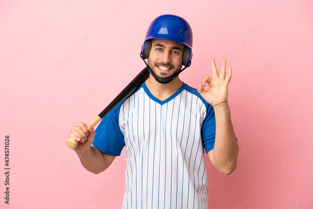Baseball player with helmet and bat isolated on pink background showing ok sign with fingers Baseball player with helmet and bat isolated on pink background showing ok sign with fingers
