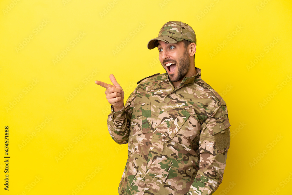 Military man isolated on yellow background pointing finger to the side and presenting a product Military man isolated on yellow background pointing finger to the side and presenting a product
