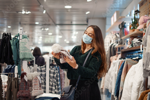 Young woman in face mask choosing warm hat in shopping mall