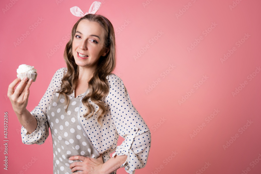 Young charming woman pastry chef and delicious muffin with cream. A cook in a polka-dot apron. pastry chef in an apron on a pink background. Young charming woman pastry chef and delicious muffin with cream. A cook in a polka-dot apron. pastry chef in an apron on a pink background.