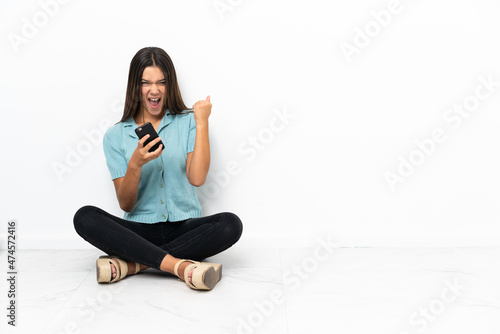 Teenager girl sitting on the floor with phone in victory position