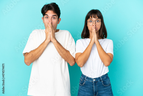Young mixed race couple isolated on blue background keeps palm together. Person asks for something