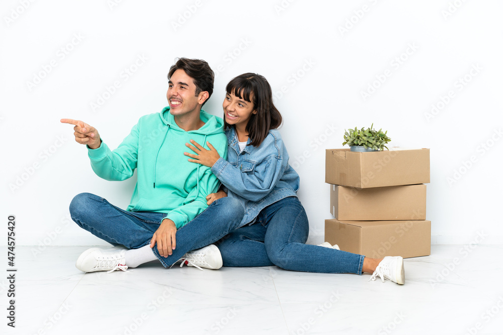 Young couple making a move while picking up a box full of things sitting on the floor isolated on white background pointing to the side to present a product Young couple making a move while picking up a box full of things sitting on the floor isolated on white background pointing to the side to present a product