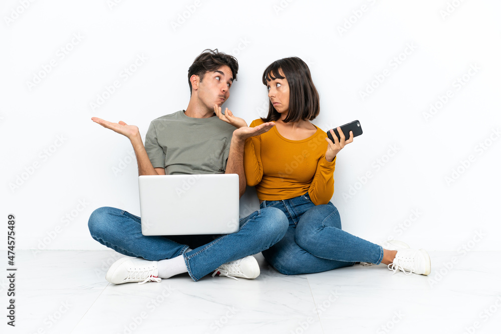 Young couple with a laptop and mobile sitting on the floor making unimportant gesture while lifting the shoulders