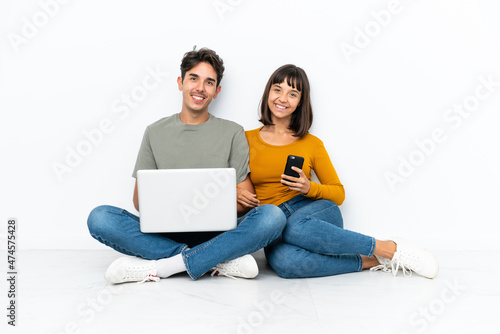 Young couple with a laptop and mobile sitting on the floor posing with arms at hip and smiling
