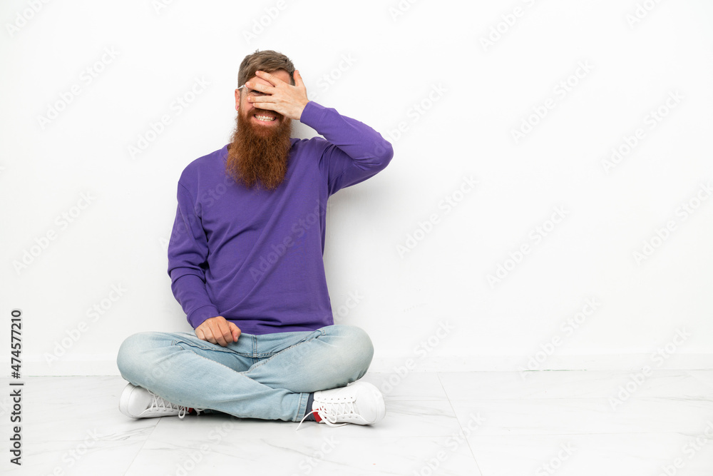 Young caucasian reddish man sitting on the floor isolated on white background covering eyes by hands and smiling Young caucasian reddish man sitting on the floor isolated on white background covering eyes by hands and smiling