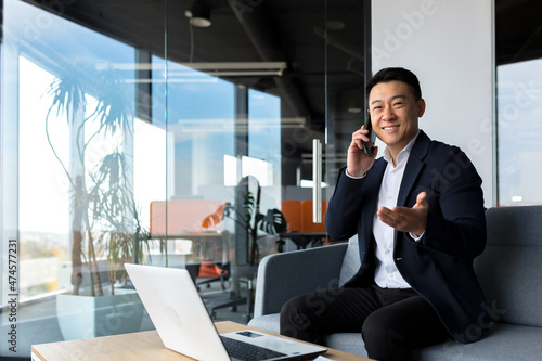 Asian businessman smiling and rejoicing happy talking on the phone, freelancer man in modern office looking at camera © Liubomir