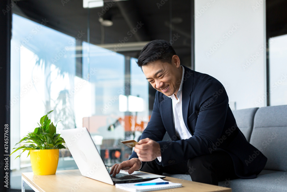 Fototapeta premium Asian boss transfers money from a bank card, sitting in the office, satisfied with the result businessman smiles, works on laptop