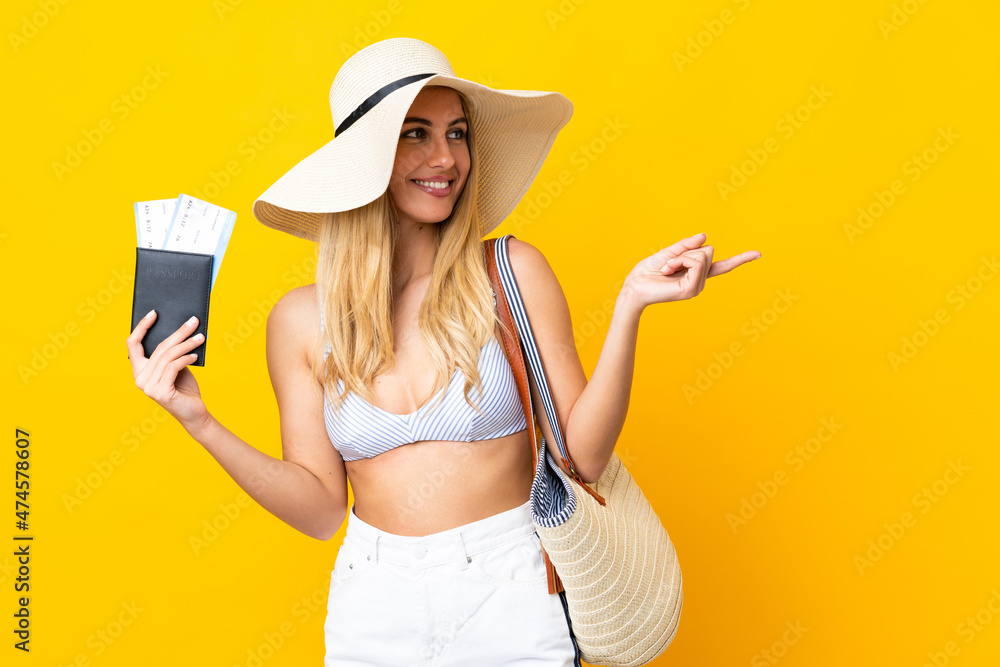 Young Uruguayan blonde woman in swimsuit holding a passport over isolated yellow background pointing to the side to present a product Young Uruguayan blonde woman in swimsuit holding a passport over isolated yellow background pointing to the side to present a product