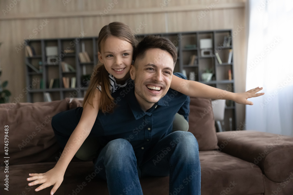 Happy young caring father holding on back adorable small kid daughter, playing airplane game sitting on cozy couch, entertaining involve in playtime weekend activity at home, single parenting concept. Happy young caring father holding on back adorable small kid daughter, playing airplane game sitting on cozy couch, entertaining involve in playtime weekend activity at home, single parenting concept.