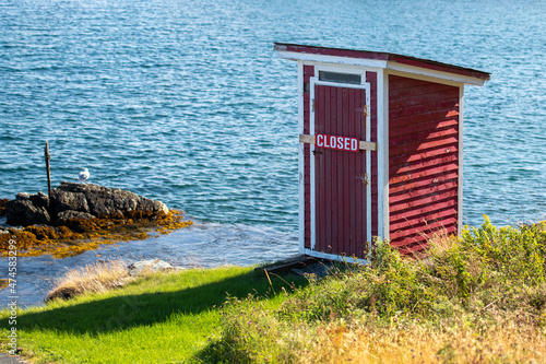 A small red building, outhouse, on the edge of a rocky cliff with blue ocean and islands in the background.  The outhouse has a closed sign across its door. The sky is blue with some white clouds.  photo