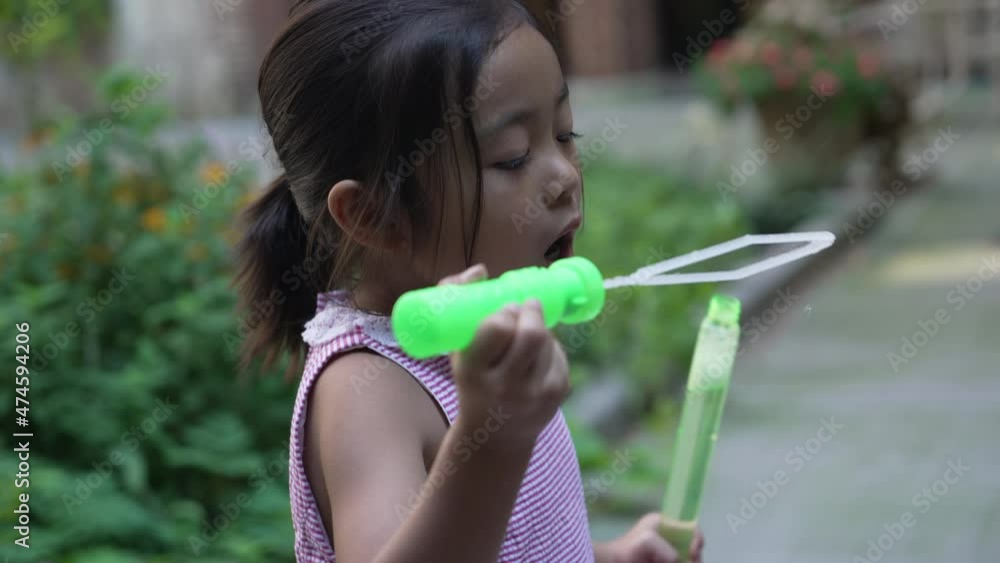 Asian female kid blowing bubbles with a green bubble wand in the courtyard Asian female kid blowing bubbles with a green bubble wand in the courtyard