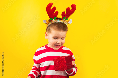 Funny kid in a reindeer costume for Christmas looks into a gift box isolated on a yellow background. Holidays, traditions, discounts, Black Friday concept. Selective focus.