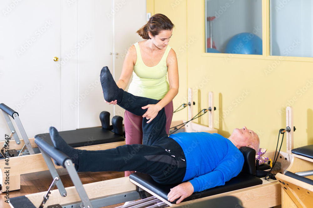 Personal female trainer controlling movements of mature man doing pilates on reformer in fitness studio.