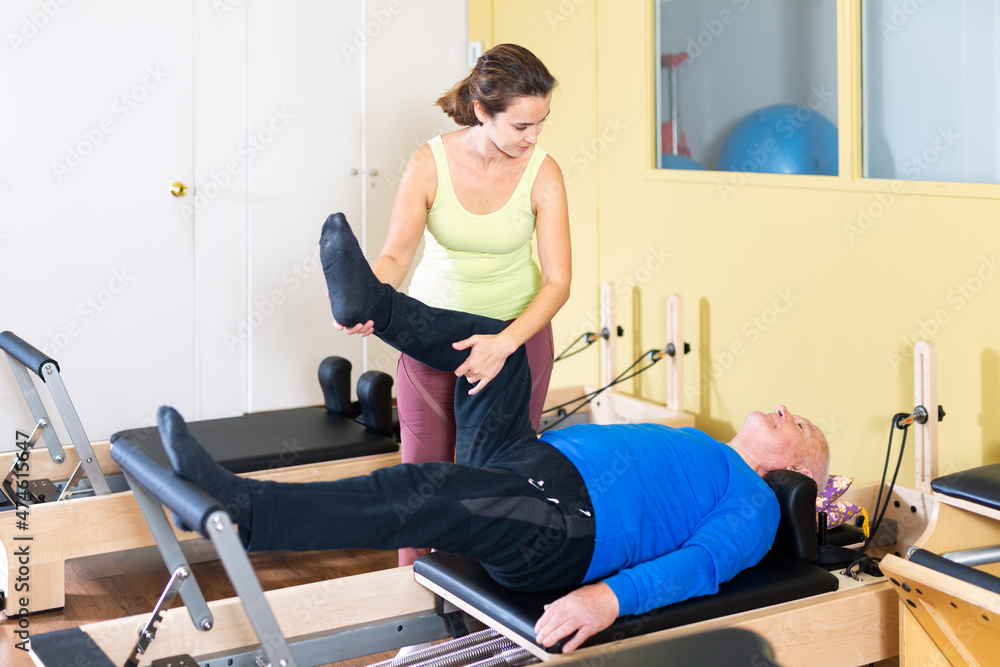 Fototapeta premium Personal female trainer controlling movements of mature man doing pilates on reformer in fitness studio.