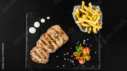 Grilled pork loin meal with fries in a basket on a black plate with sauces and fresh veggies