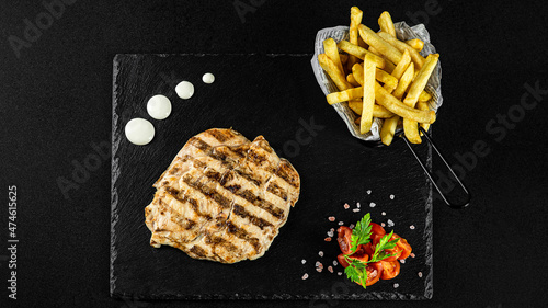Grilled Chicken breast  meal with fries in a basket on a black plate with sauces and fresh veggies