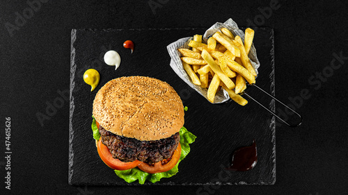 Grilled spicy Gurmanska Pleskavica Burger with cheese, fries in a basket on a black plate with sauces and fresh veggies photo