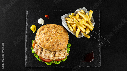 Grilled Chicken breast Burger with fries in a basket on a black plate with sauces and fresh veggies