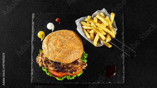 Grilled Pork Steak Burger with fries in a basket on a black plate with sauces and fresh veggies