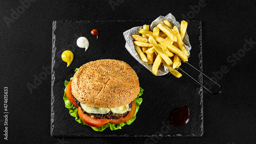 Grilled Gurmanska Pleskavica Burger with cheese,fries in a basket on a black plate with sauces and fresh veggies