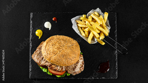 Grilled Pork Loin Burger with fries in a basket on a black plate with sauces and fresh veggies