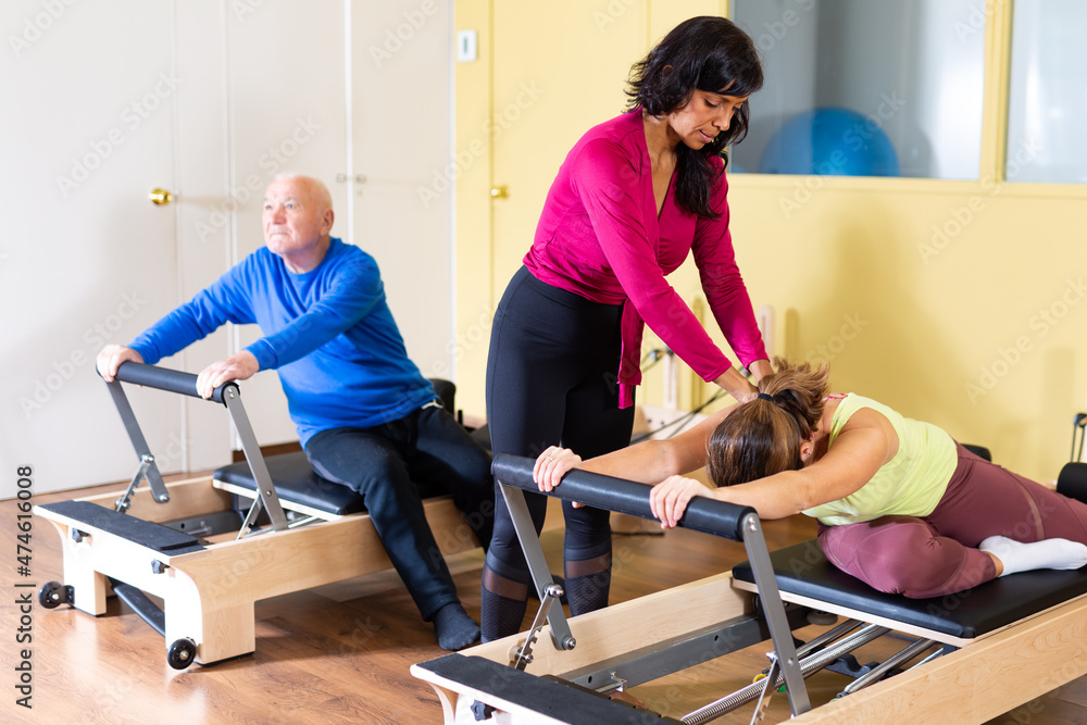 Fototapeta premium Hispanic female pilates instructor helping young woman doing exercises on reformer apparatus at gym..
